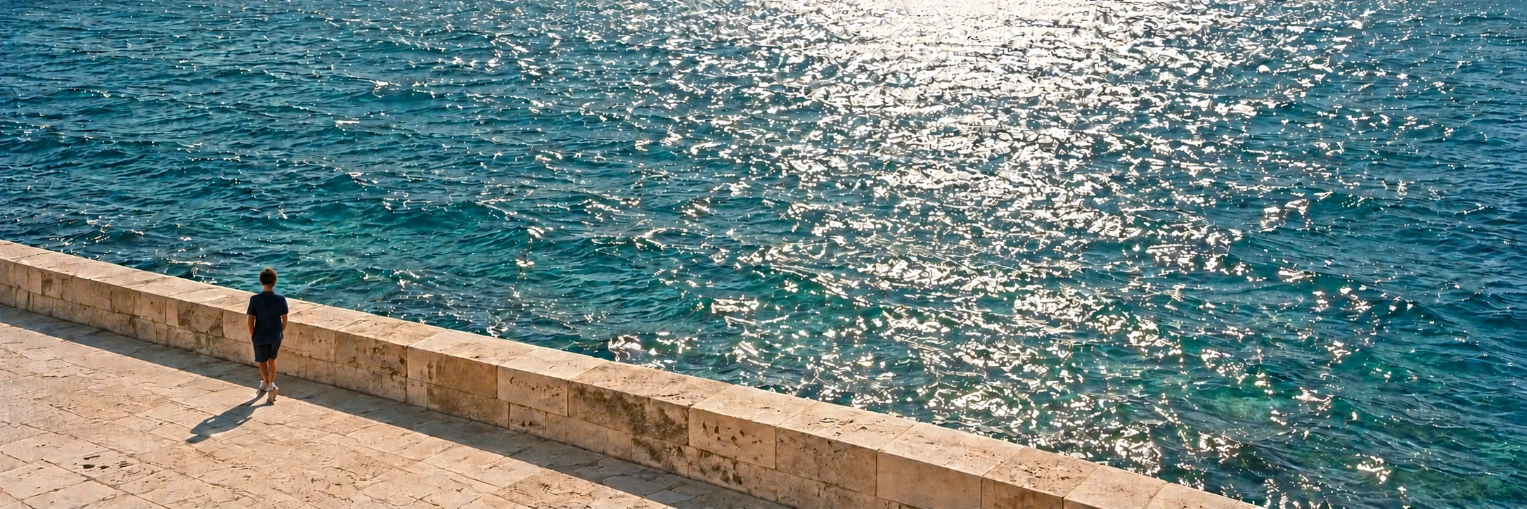 A small figure seen from behind, walking along the limestone seafront beside sunlit, shimmering water.